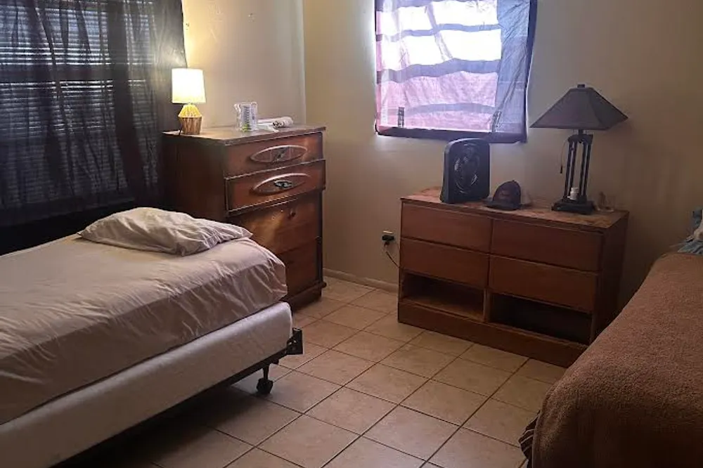 A modestly furnished inpatient housing room featuring a single bed, wooden dressers, a lamp, and tiled flooring.